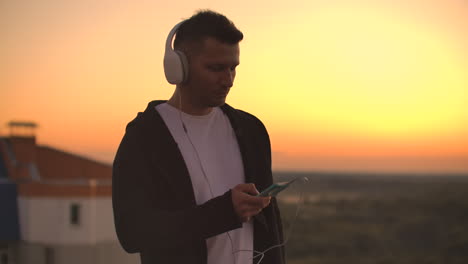 A-man-in-big-white-headphones-listens-to-music-standing-on-the-roof-at-sunset.-Relaxation.